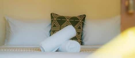 A bed with white bedding, a green patterned pillow, and a rolled white towel.