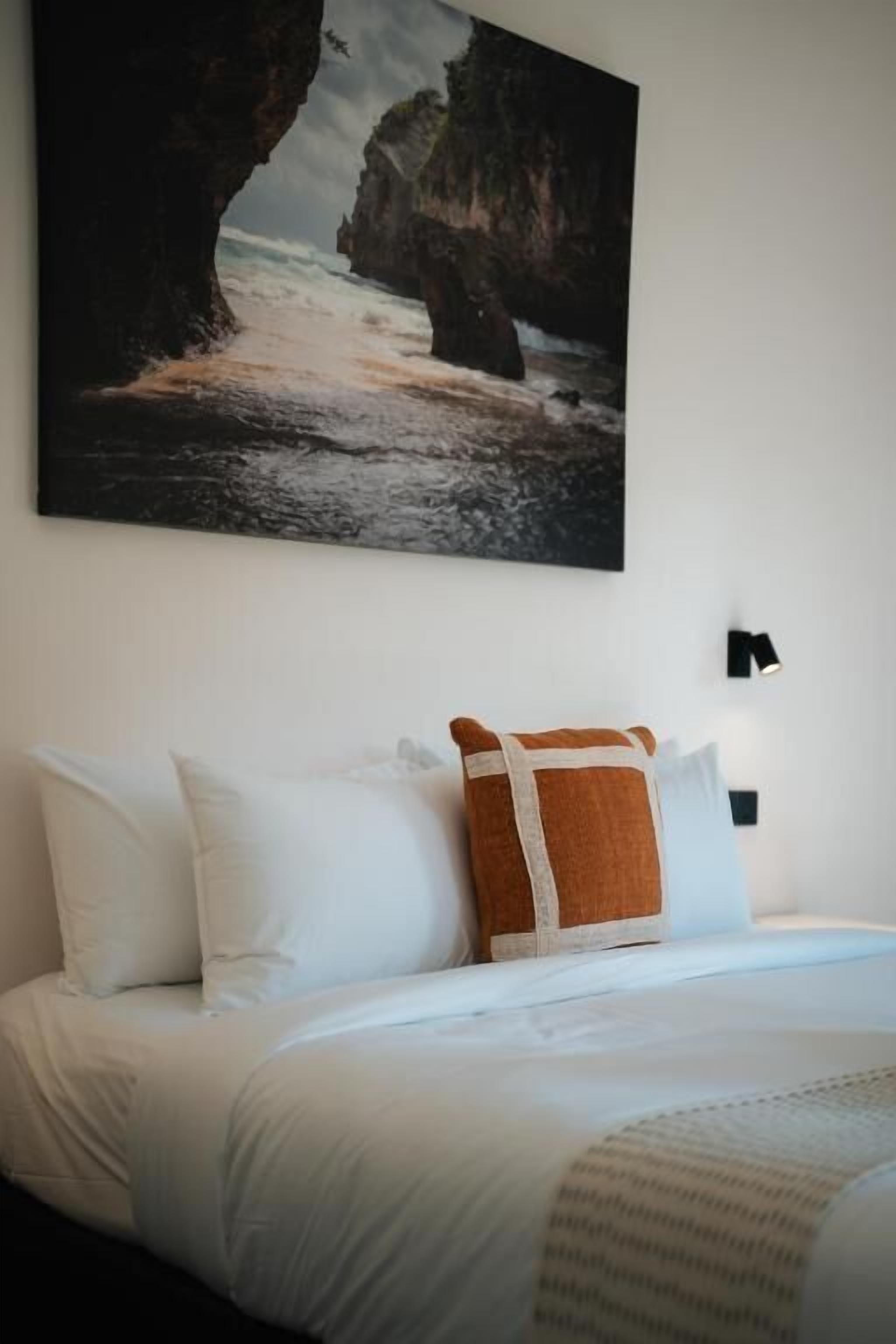 A bedroom with a bed, pillows, a blanket, a painting of a coastal scene, and a wall-mounted light.