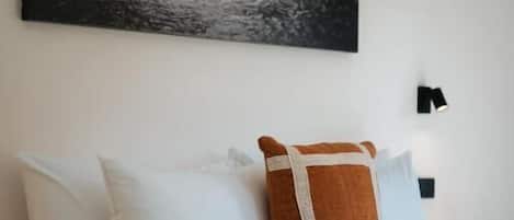 A bedroom with a bed, pillows, a blanket, a painting of a coastal scene, and a wall-mounted light.