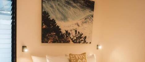 A bedroom with a bed, bedside tables, a painting, and an air conditioning unit.