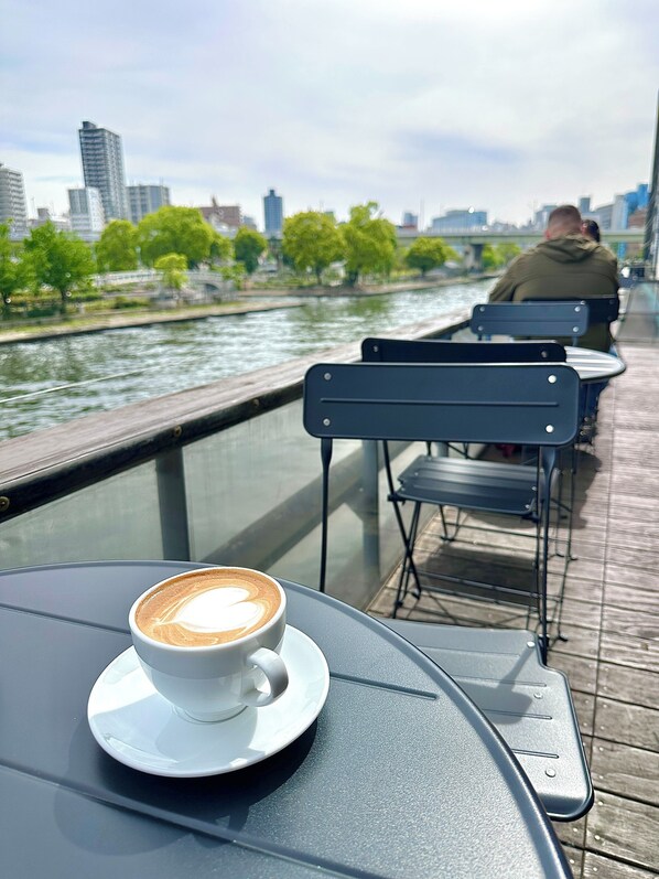 Ein Tisch mit einer Kaffeetasse, einem Stuhl und Blick auf einen Fluss mit Gebäuden im Hintergrund.