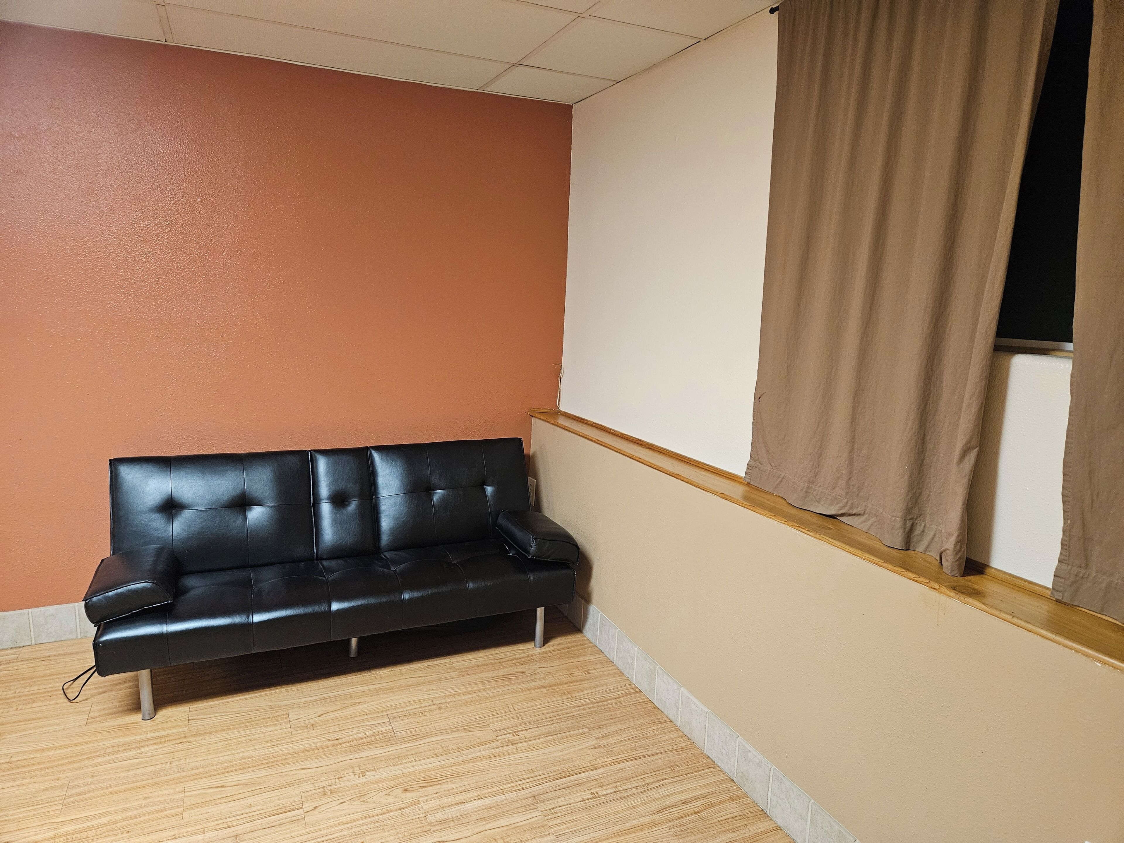 A black leather sofa in a corner with a wooden floor, a beige wall, and a window with a brown curtain.