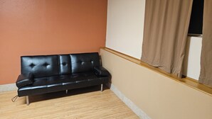 Un divano in pelle nera in un angolo, con pavimento in legno, parete color crema e una finestra con tenda marrone.