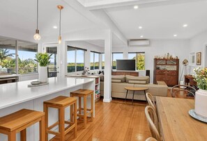 A modern kitchen with a large island, wooden stools, and a dining table. - SaltNest - A calm and modern retreat by the sea (Bundeena)