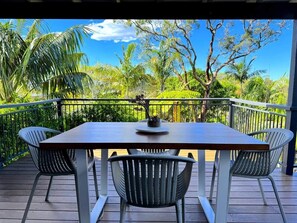 A wooden deck with a table and chairs, surrounded by lush greenery and a clear blue sky. - SaltNest - A calm and modern retreat by the sea (Bundeena)