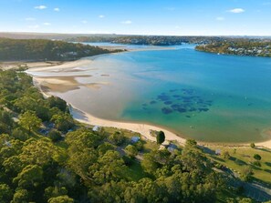 An aerial view of a coastal area with a beach, clear waters, and surrounding greenery. - SaltNest - A calm and modern retreat by the sea (Bundeena)