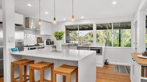 Una cocina moderna con armarios blancos, una isla grande con taburetes, un dispensador de agua y vistas a zonas verdes a través de amplios ventanales.