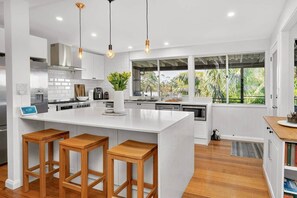 Una cocina moderna con armarios blancos, una isla grande con taburetes, un dispensador de agua y vistas a zonas verdes a través de amplios ventanales.