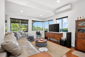 A spacious living room with a leather sofa, a round coffee table, a TV on a wooden cabinet, and large windows offering a view of greenery. - SaltNest - A calm and modern retreat by the sea (Bundeena)