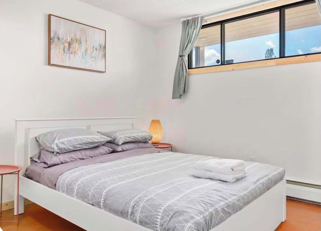Un dormitorio con cama, mesita de noche, lámpara, ventana con cortinas y un cuadro en la pared.