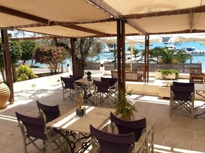 An outdoor patio with tables and chairs, a pergola, and a view of boats on the water. - HOTEL ROZOS (Ermionida)