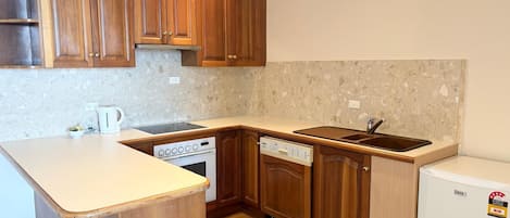 A compact kitchen with wooden cabinets, a built-in dishwasher, a small refrigerator, and a countertop with a kettle.