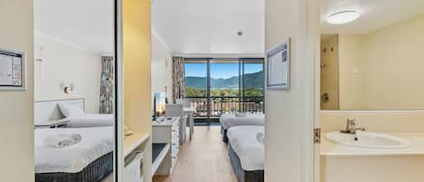 A hotel room with two beds, a desk, a chair, a large window with a mountain view, and a bathroom with a sink and mirror.