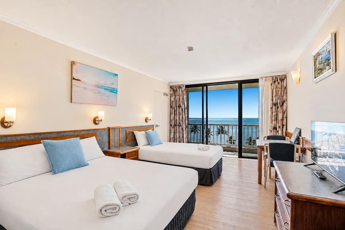 A hotel room with two beds, a desk, a TV, and a balcony with a view of the sea.