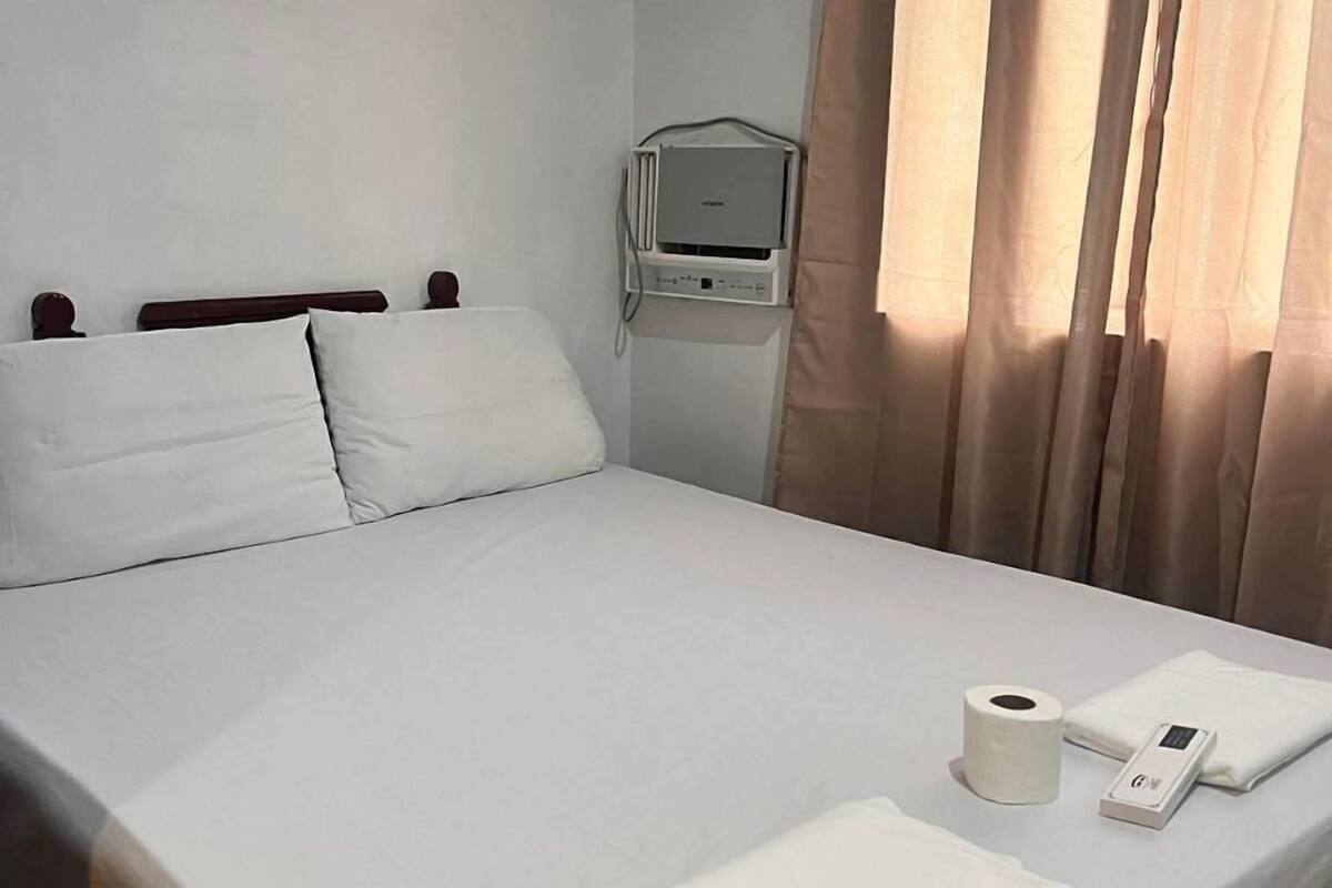 Una cama bien hecha con ropa blanca, una mesita de noche con control remoto, un rollo de papel higiénico y un juego de toallas.