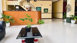 Vestíbulo de hotel con mostrador de recepción, escalera y plantas en macetas.