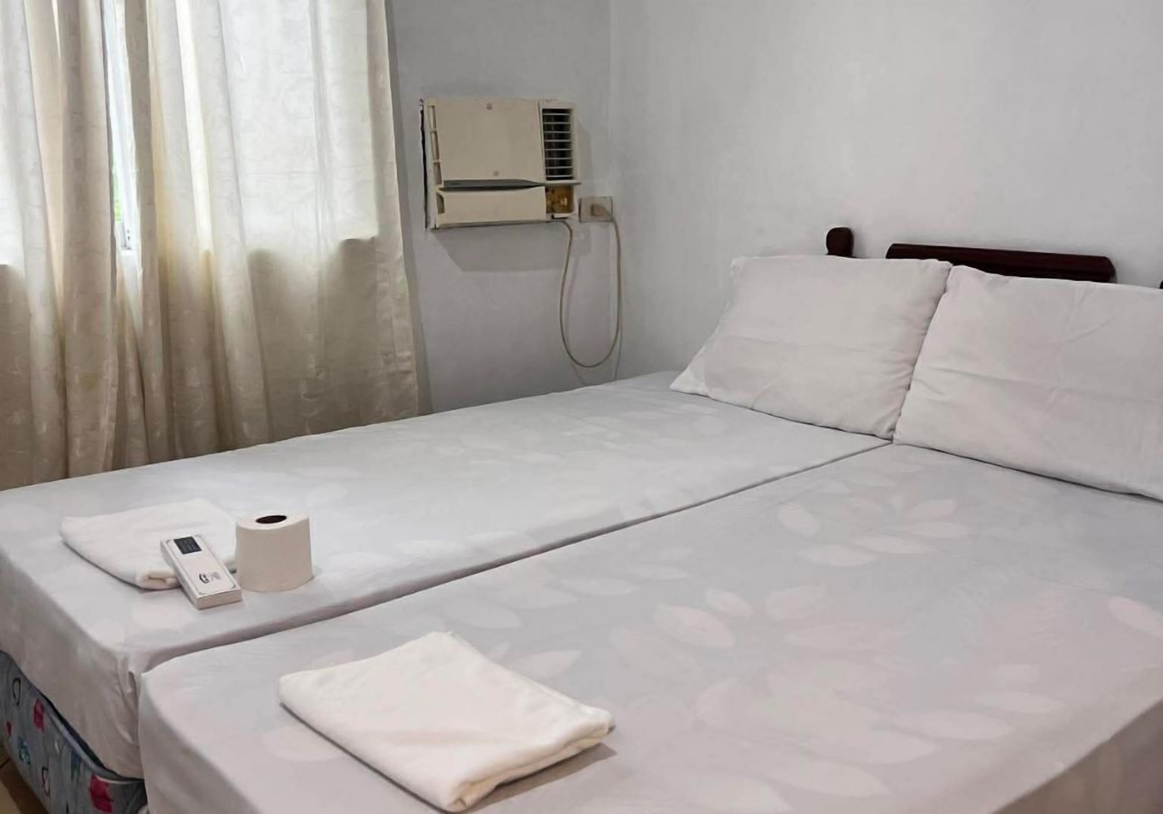 A hotel room with a bed, a bedside table with a remote control and a tissue box, a wall-mounted air conditioning unit, and a window with curtains.