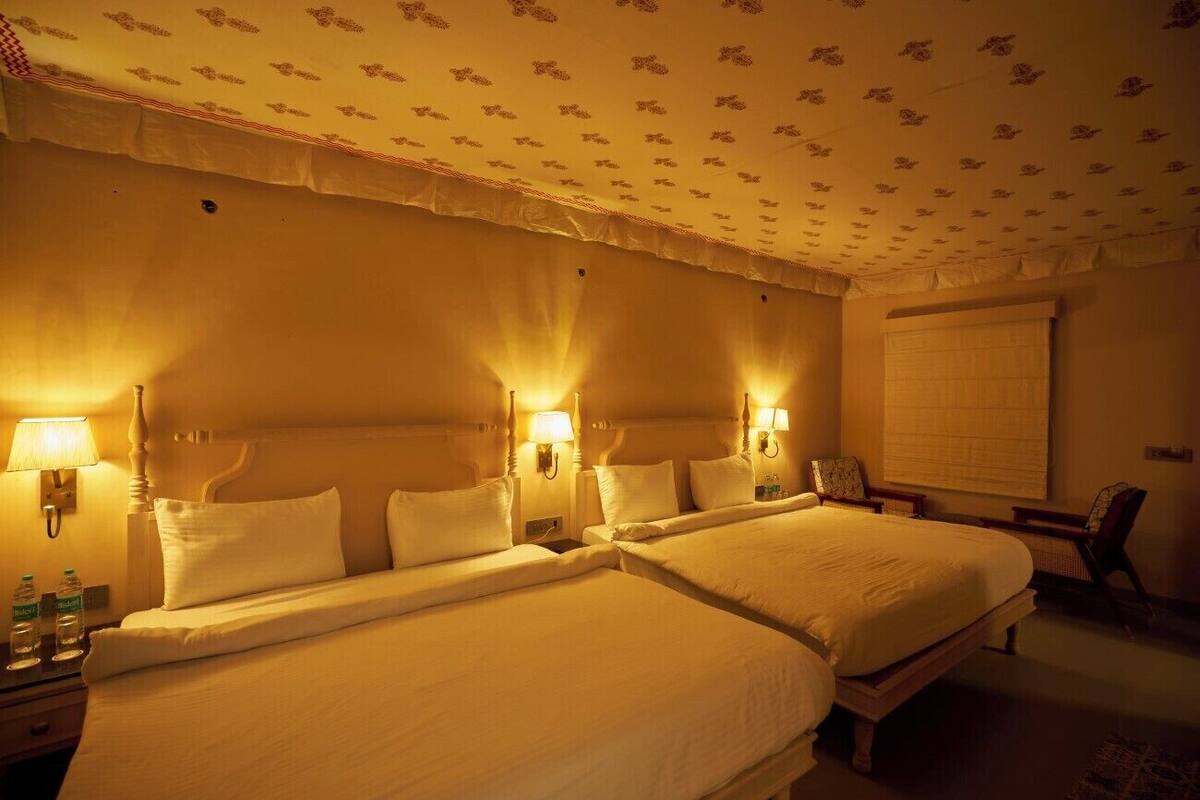Habitación de hotel con dos camas, lámparas fijadas a la pared, una silla y una ventana con persianas.