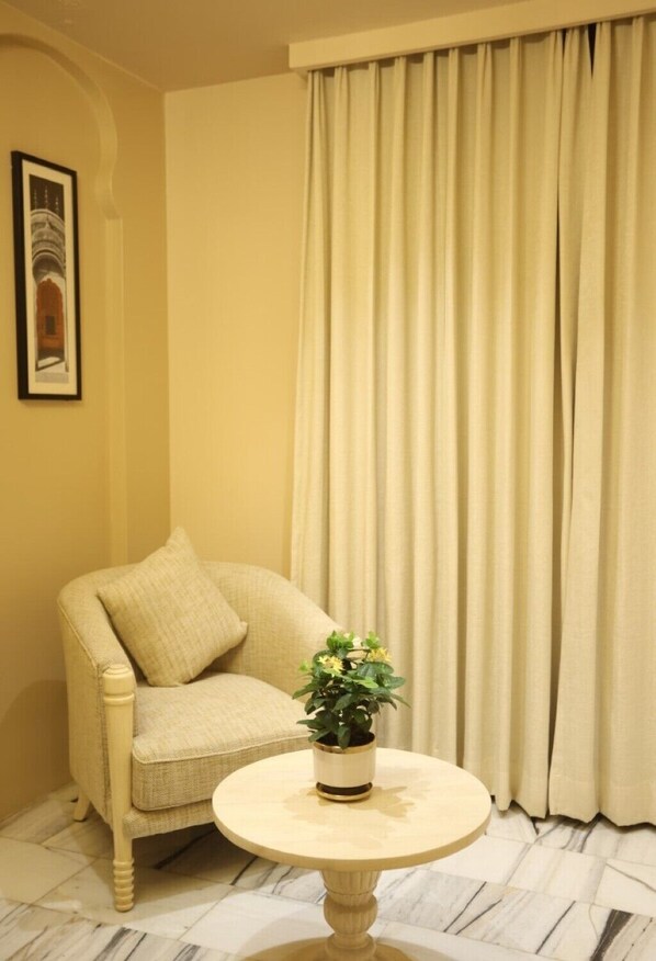 A room with a beige armchair, a small round table with a potted plant, a framed picture on the wall, and a large curtain.
