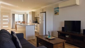 Un salon moderne avec un canapé, une table basse en bois, un téléviseur posé sur un meuble et une cuisine en arrière-plan.
