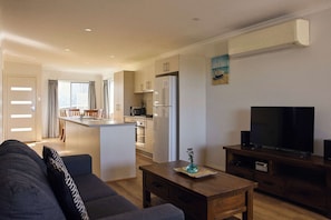 A modern living room with a sofa, a wooden coffee table, a TV on a stand, and a kitchen in the background. - Currowan House - Three-Bedroom Home with Balcony ( (Mollymook)