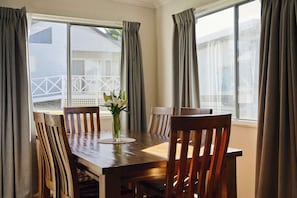 A dining table with chairs, a vase with flowers, and curtains on windows. - Currowan House - Three-Bedroom Home with Balcony ( (Mollymook)