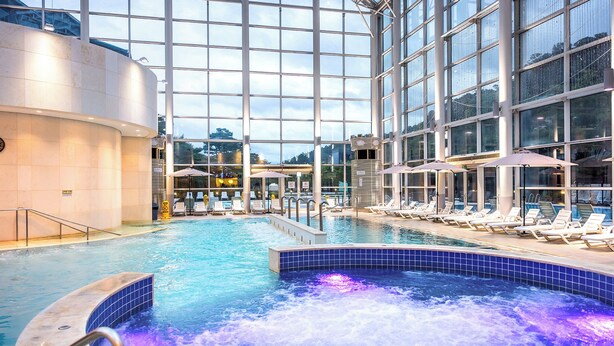 Een binnenzwembad met ligbedden en een jacuzzi, omgeven door grote ramen.