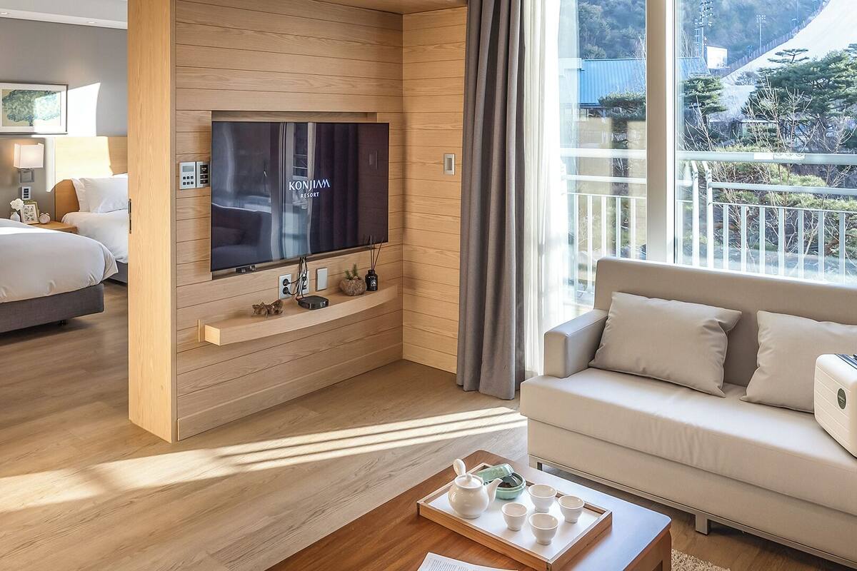 A hotel room with a bed, a sofa, a TV mounted on a wooden wall, and a balcony with a view.