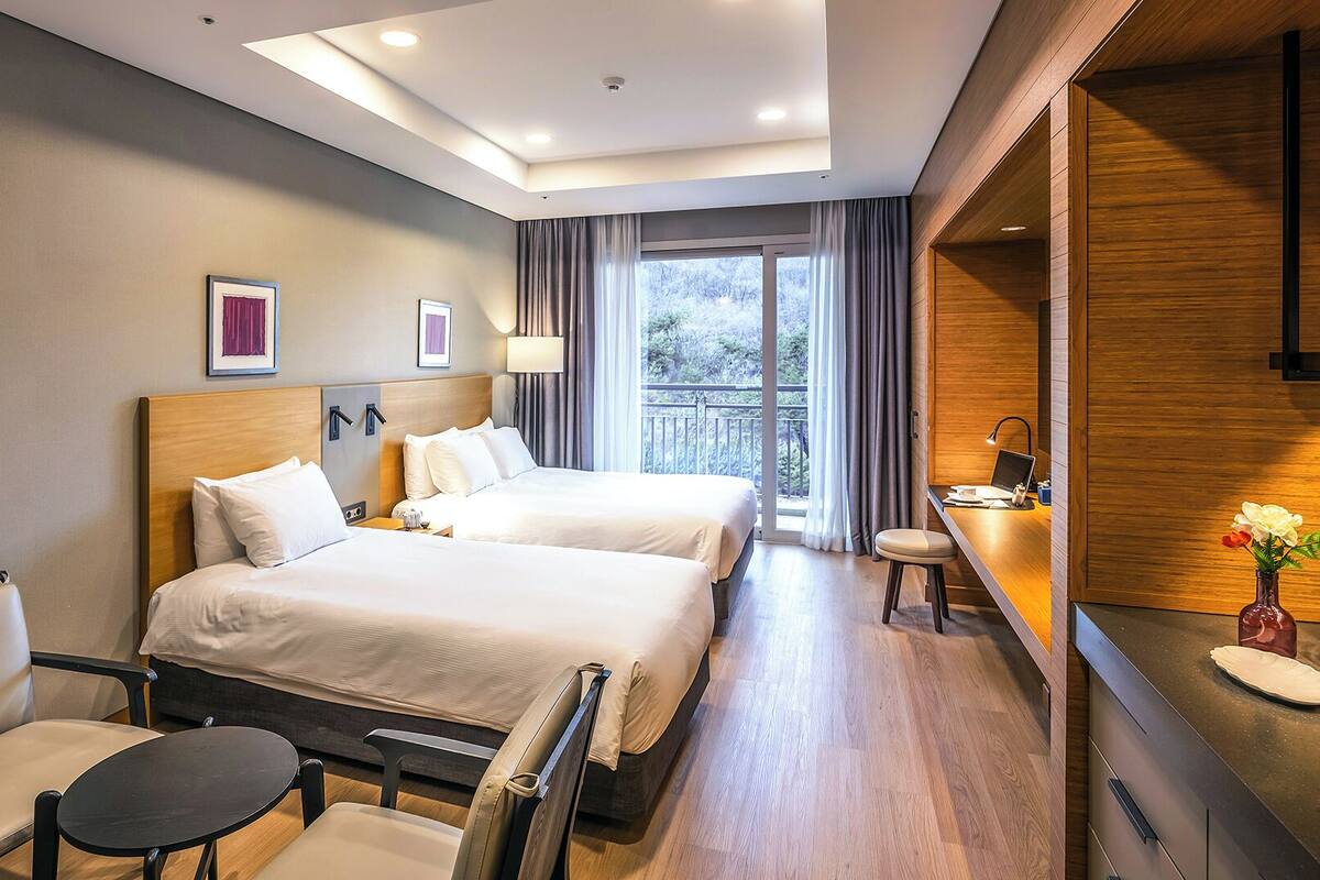 A hotel room with two beds, a desk, a chair, and a view of the outdoors.