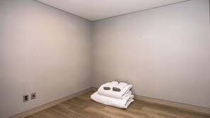 A corner room with light-colored walls, a wooden floor, and a stack of white bedding.