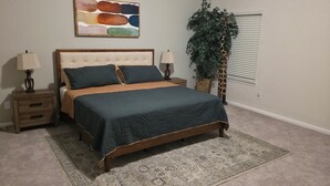 A bedroom with a bed, nightstands, lamps, a large plant, and a wall art piece.
