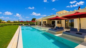 Une piscine avec des chaises longues et des parasols rouges, un espace extérieur aménagé et abrité, et un ciel d’un bleu limpide.