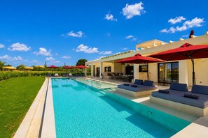 A swimming pool with lounge chairs and red umbrellas, a covered outdoor seating area, and a clear blue sky. - Villa Nobilita - Luxury villa in Corfu. At the beach, roof terrace and pool (Corfu)