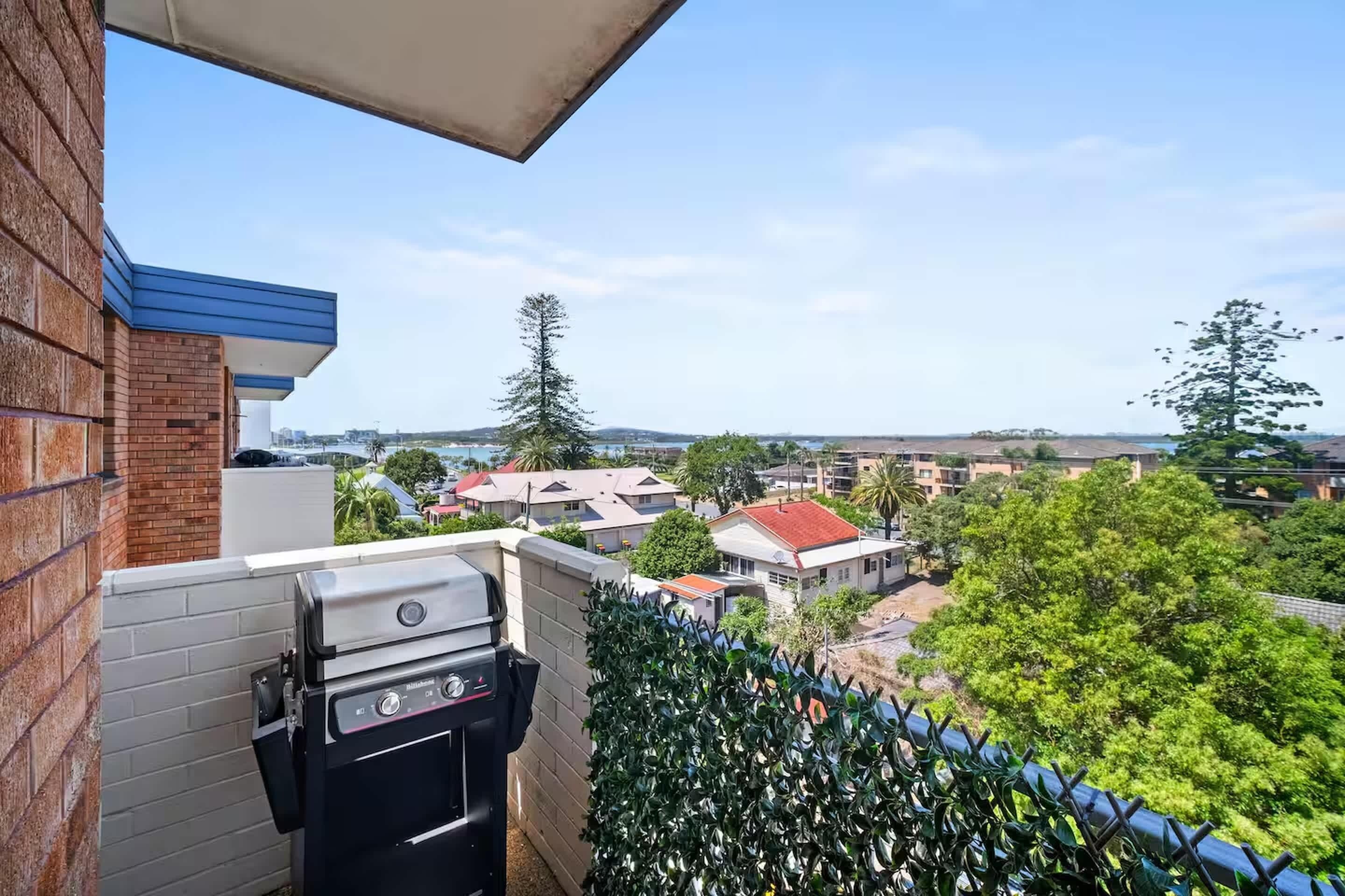 A balcony with a barbecue grill, a view of a residential area, and a hedge.