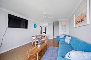 Ein modernes Wohnzimmer mit Couchtisch aus Holz, einem blauen Sofa, einem Esstisch mit Stühlen und einem Deckenventilator.