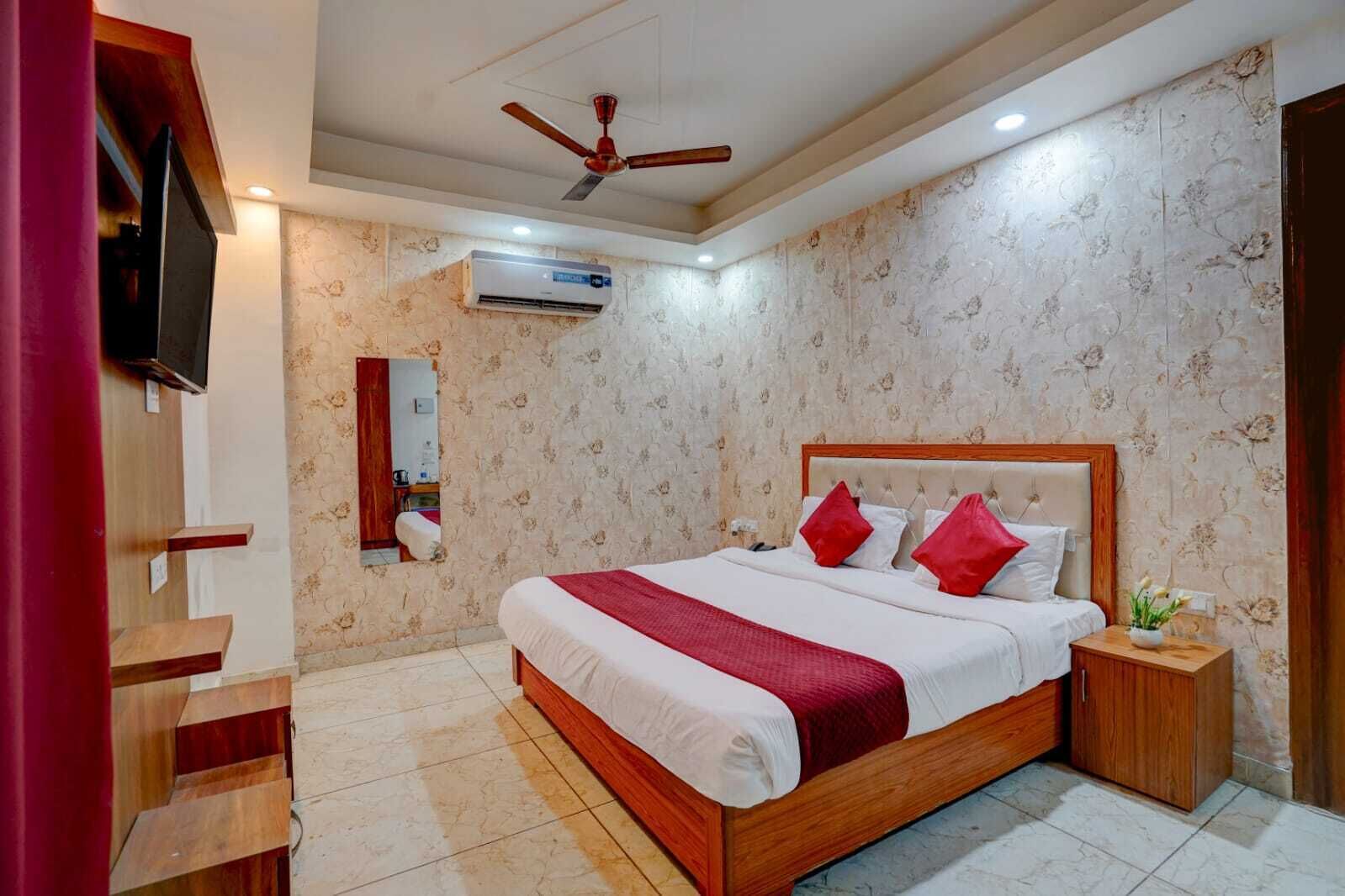 A bedroom with a wooden bed, red pillows, a ceiling fan, an air conditioning unit, a TV, and a mirror.