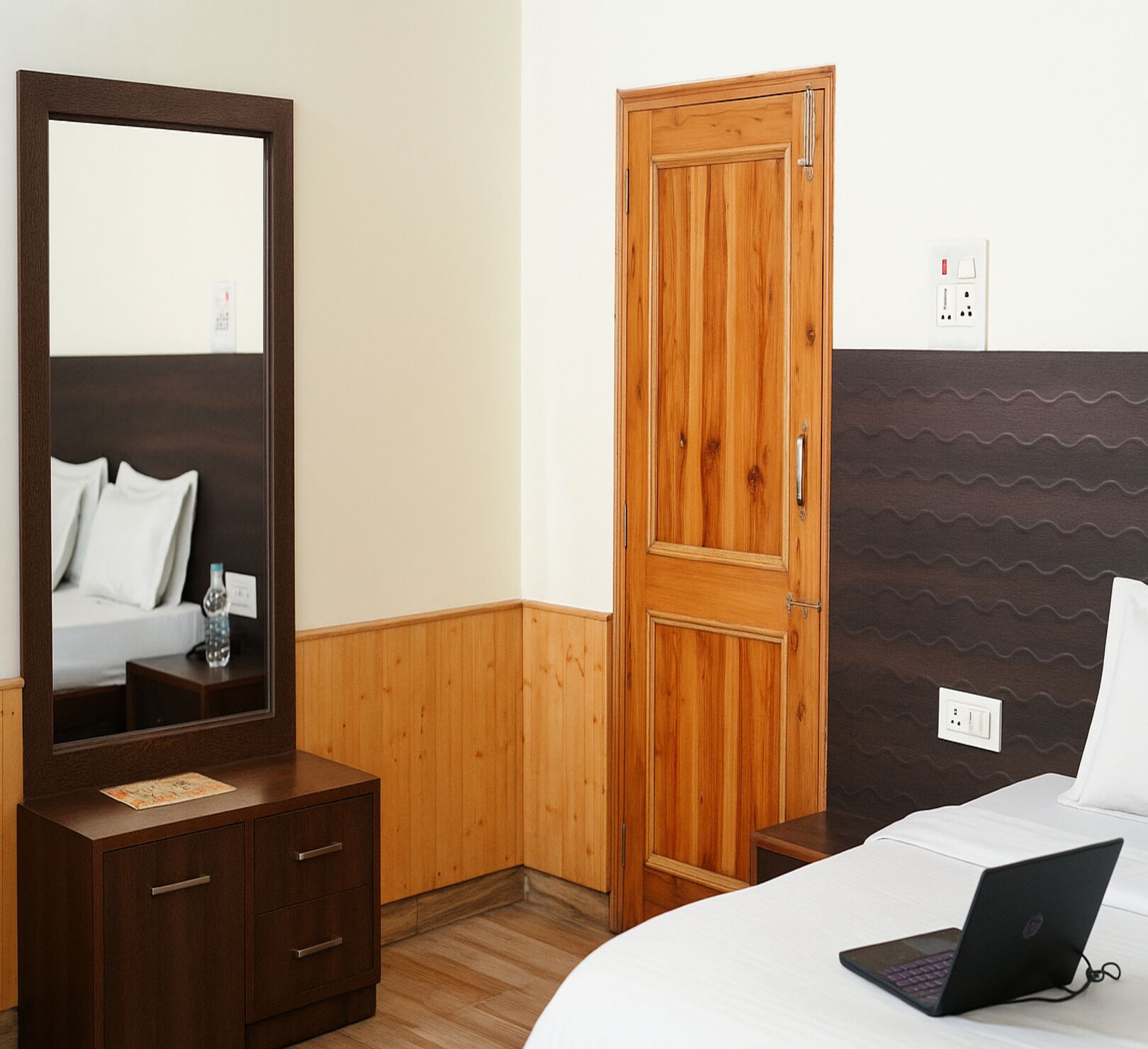 A hotel room with a wooden wardrobe, a large mirror, a bed with a laptop, and a wall-mounted electrical outlet.