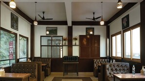 A restaurant with booths and tables, ceiling fans, pendant lights, and large windows. - Chaman Vatika (Kasol)