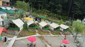 A cluster of small, green-roofed cabins with outdoor seating areas, surrounded by a stone wall and greenery. - Chaman Vatika (Kasol)