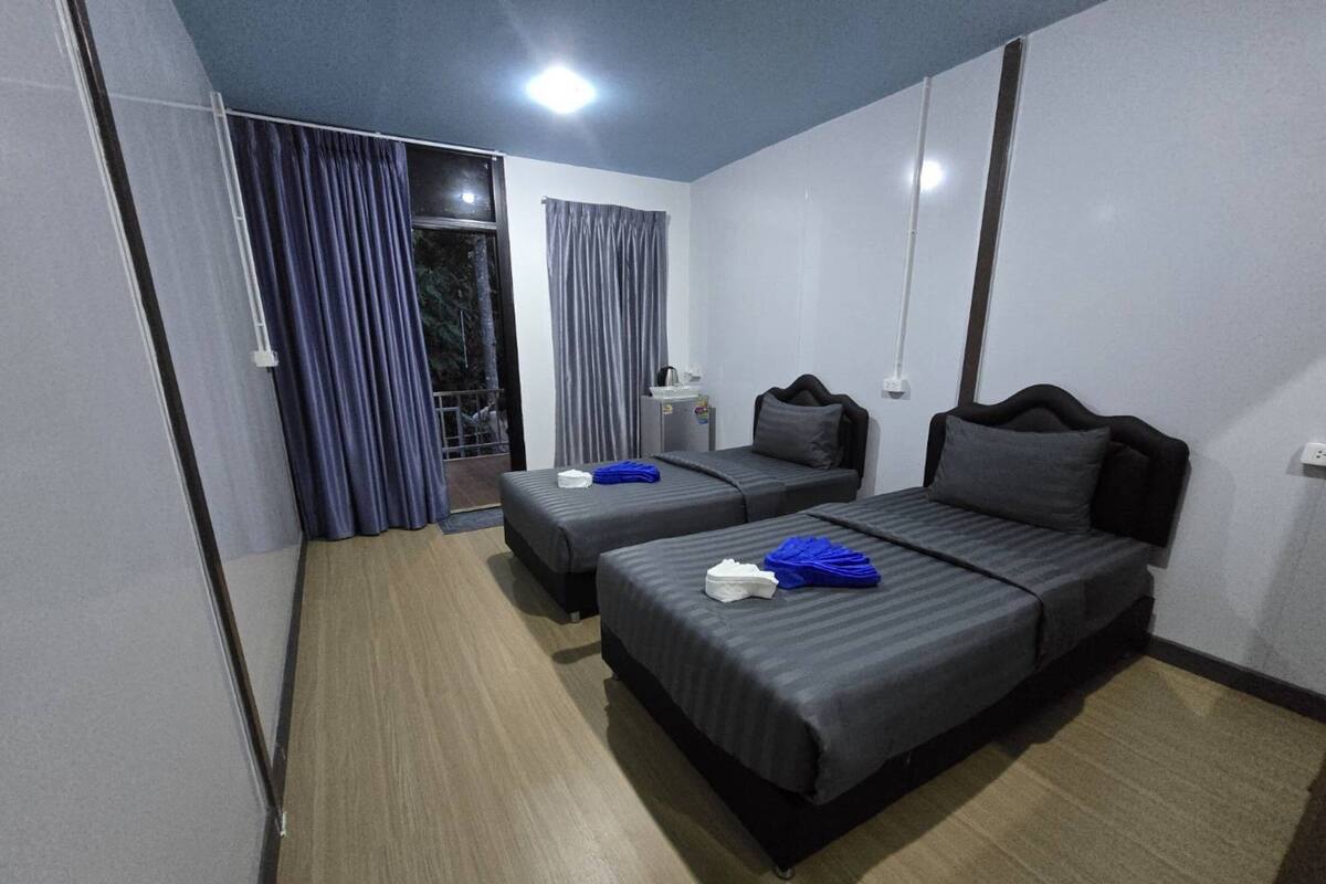 A hotel room with two beds, a small table, a window with curtains, and a door with a metal gate.