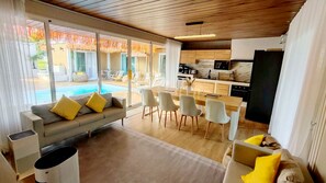 En moderne stue med sofa, spisebordsstole, et tekøkken og udsigt til en pool.