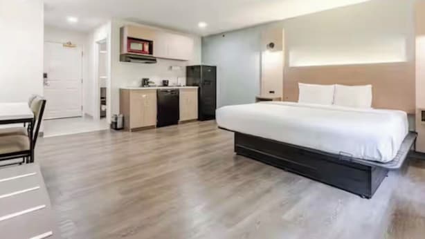 A modern hotel room with a large bed, a kitchenette, and a dining area.