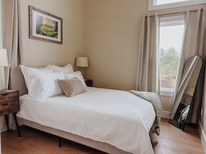 A neatly made bed with white linens, a bedside table with a lamp, a framed picture on the wall, and a window with curtains. - Peaceful one-bedroom mountain retreat in Floyd, Virginia featuring a full kitchen, cozy living space, private outdoor seating, and shared fire pit access just minutes from downtown Floyd. (Floyd)