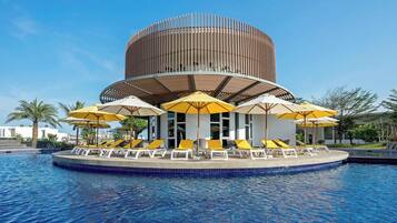 A circular pool with lounge chairs and umbrellas, a modern building with vertical slats, and palm trees in the background.