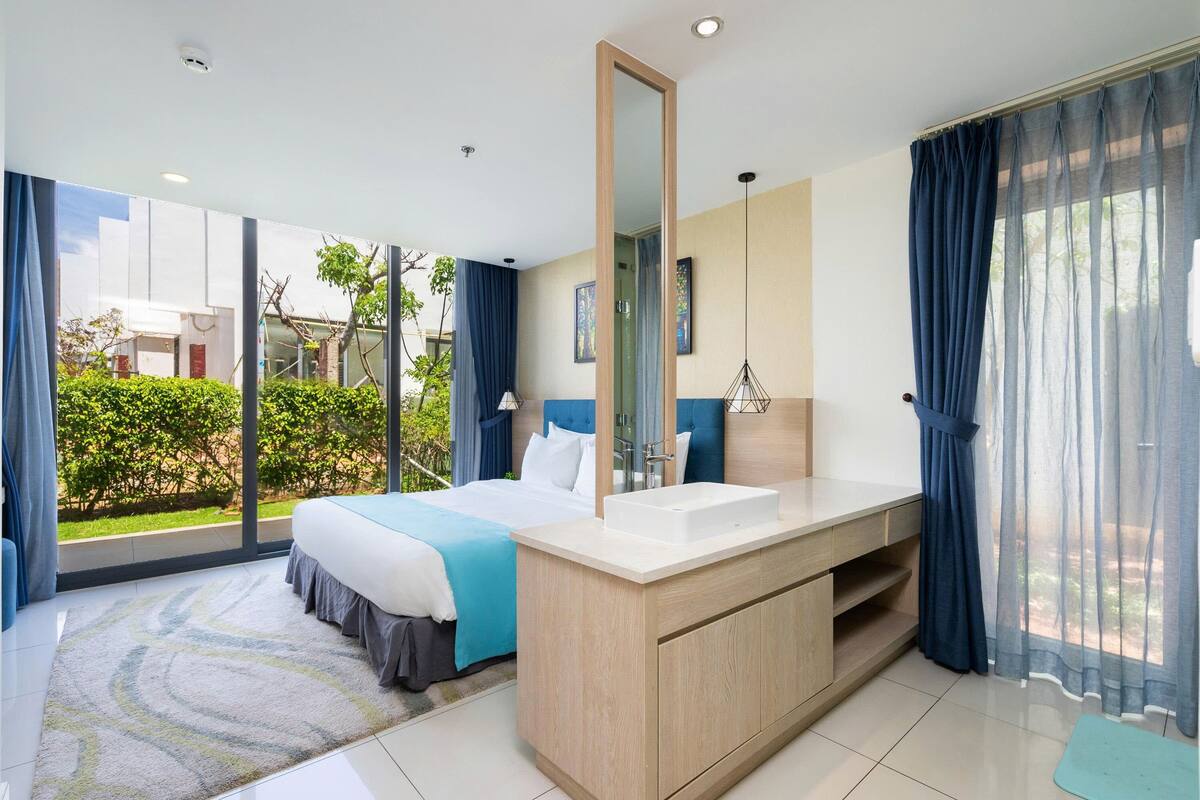 A modern hotel room with a large bed, a bathroom with a vanity, and a view of greenery outside.