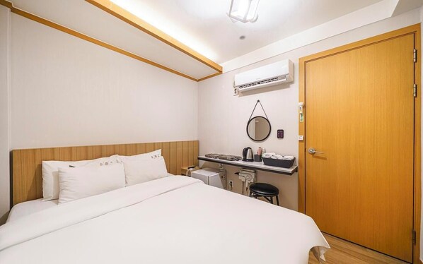 A hotel room with a bed, a desk with a chair, an air conditioning unit, a door, and a mirror on the wall.