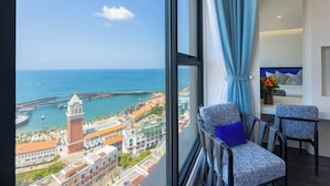 Habitación de hotel con un ventanal grande que ofrece vistas a una ciudad costera, incluyendo una torre de reloj destacada y un puente.