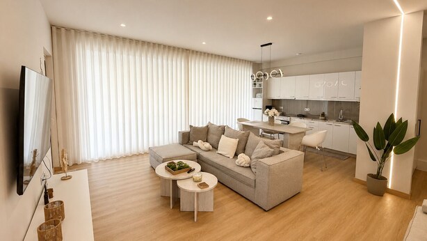 A modern living room with a sofa, coffee table, and a TV. There's a kitchen in the background with white cabinets and a dining area. - Akrata Comfort Living (Krathi)