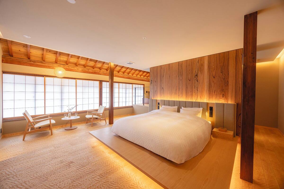 A modern bedroom with a large bed, wooden accents, a small table with chairs, and large windows.