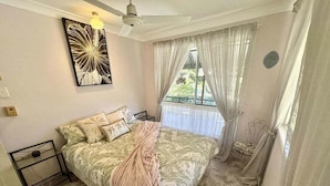 Une chambre à coucher comprenant un lit, un ventilateur de plafond, une fenêtre avec des rideaux et une œuvre d’art accrochée au mur.